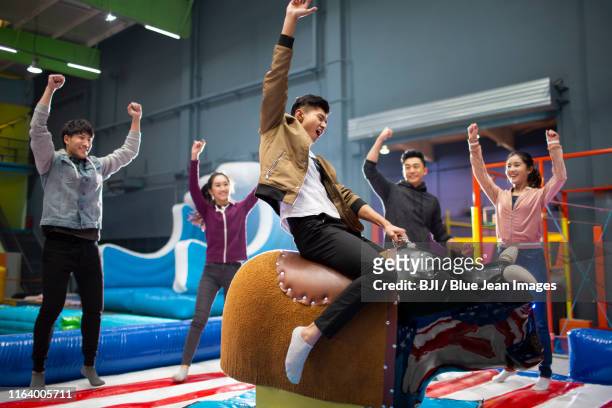 happy young chinese friends playing mechanical bull game - bucking bronco stock-fotos und bilder