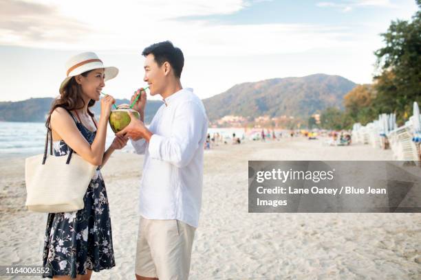 happy young chinese couple drinking coconut milk on beach - 2 coconut drinks ストックフォトと画像