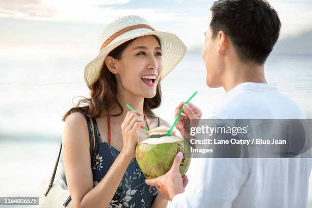 happy young chinese couple drinking coconut milk on beach - acqua di cocco foto e immagini stock