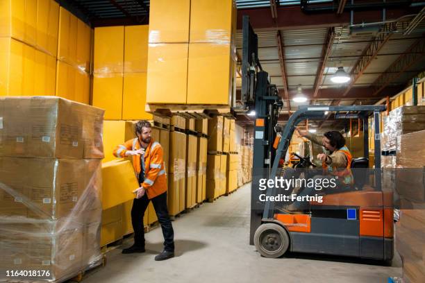 an industrial warehouse workplace safety topic. a worker in the danger zone, walking close to a raised forklift carrying a load. - workplace danger stock pictures, royalty-free photos & images
