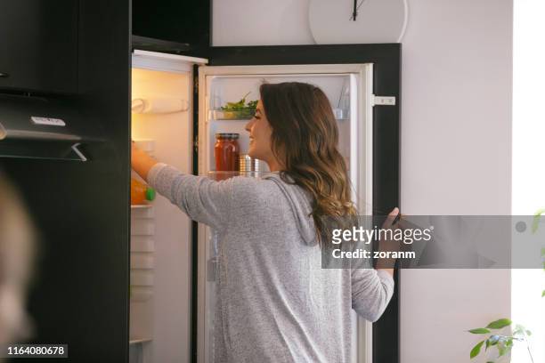 glückliche frau, die lebensmittel aus dem kühlschrank nimmt - refrigerator stock-fotos und bilder