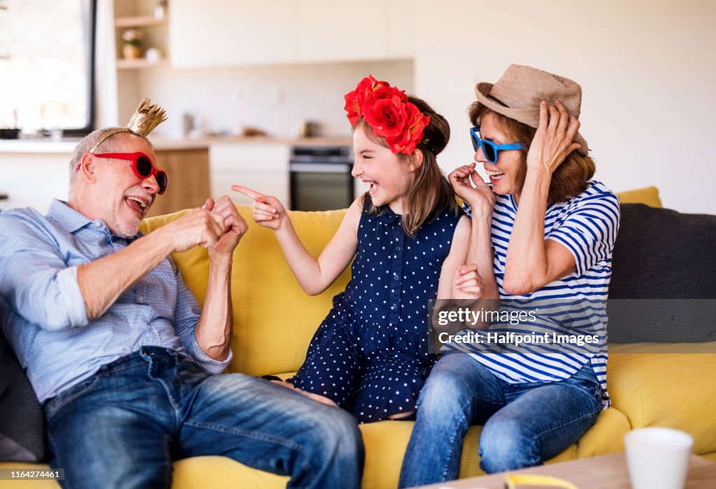 Senior grandparents and granddaughter sitting indoors on sofa, having fun.