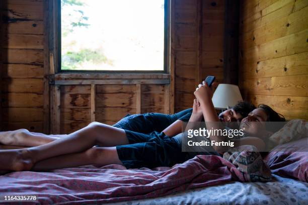 relaxing at the summer cabin.  two brothers relax on a bed with a smart phone in the bedroom of a rustic summer cabin or cottage. - cabin stock pictures, royalty-free photos & images