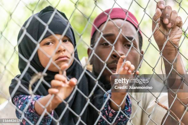 muslim mid adult black man holding his daughter looking through a fence - refugee camp stock pictures, royalty-free photos & images