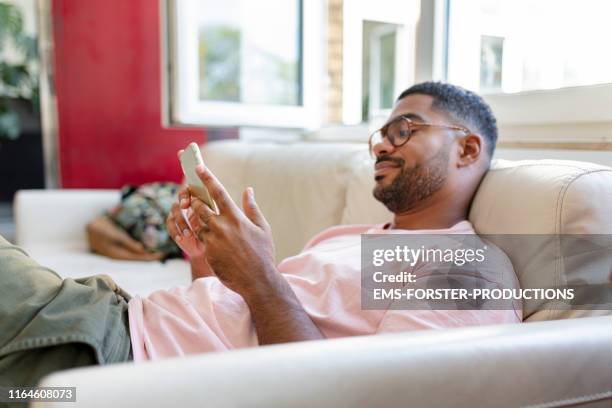 relaxed man sitting on sofa using cell phone - male looking content stockfoto's en -beelden