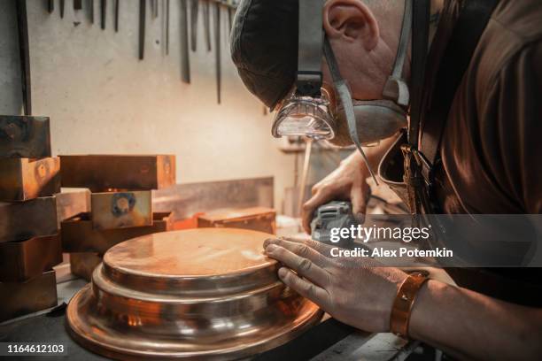 el artesano que lleva gafas protectoras y el delantal de cuero puliendo los detalles de latón con la herramienta eléctrica - abrillantar fotografías e imágenes de stock