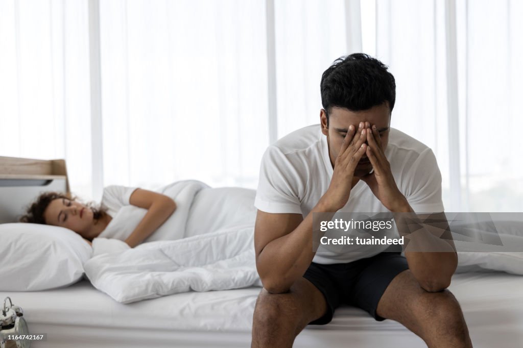 Un joven deprimido deprimido sentado en el borde de la cama contra su esposa acostada en la cama. Dificultades de relación.