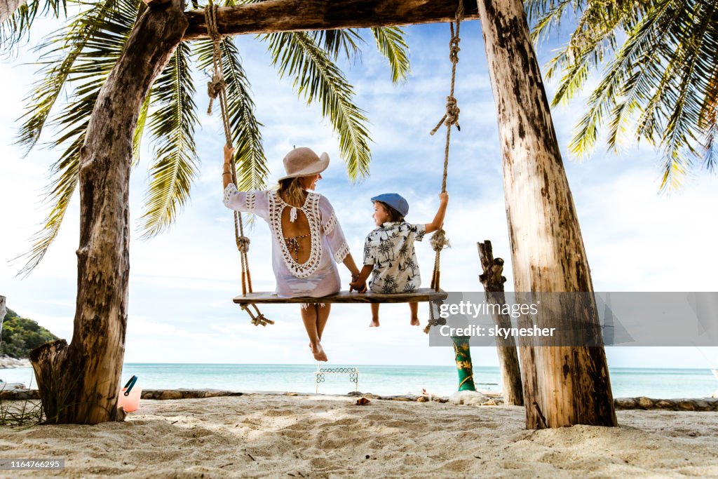 Vista trasera de madre e hijo despreocupados cogidos de la mano mientras se balancea en la playa.