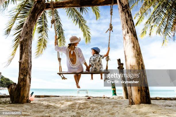 rear view of carefree mother and son holding hands while swinging at the beach. - tropical climate stock pictures, royalty-free photos & images