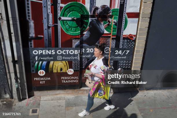 Woman smokes a cigarette as she walks past an ad for a forthcoming easyGym in Camberwell, south London, on 29th August 2019, in London, England.