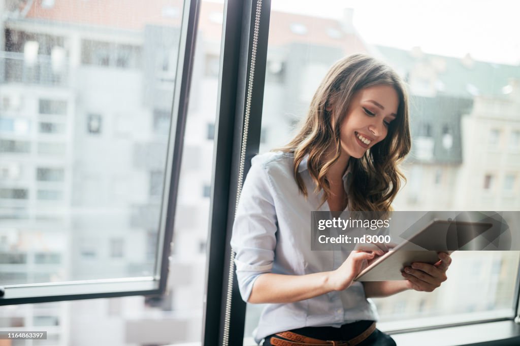 Atractiva empresaria usando una tableta digital mientras está de pie frente a las ventanas