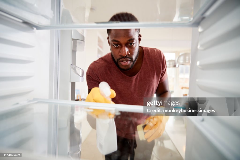 View Looking Out From Inside Empty Refrigerator As Man Wearing Rubber Gloves Cleans Shelves