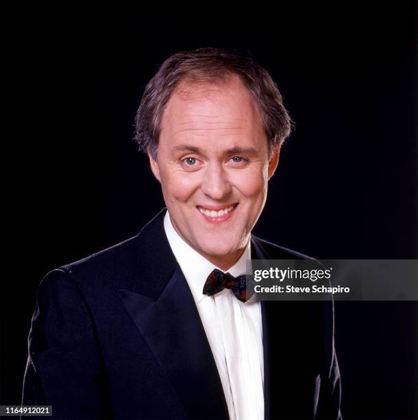 Portrait of American actor John Lithgow, dressed in a tuxedo, as he smiles and poses against a black backgorund, 1980s.