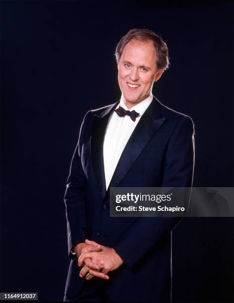 Portrait of American actor John Lithgow, dressed in a tuxedo, as he smiles and poses against a black backgorund, 1980s.