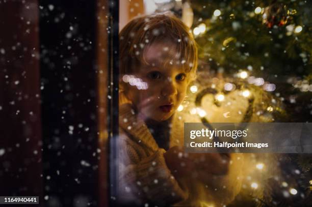 a small cute boy indoors standing by the window at christmas time, looking out. - an evening with heart fotografías e imágenes de stock