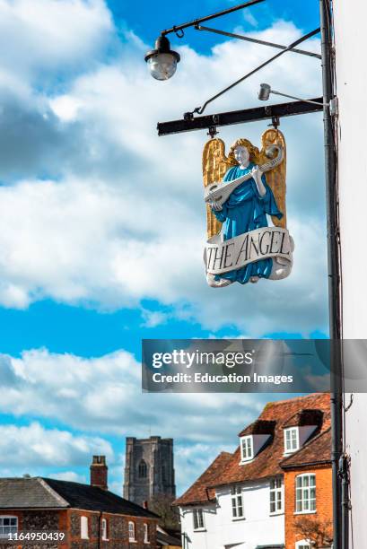 The Angel pub sign in front of the Market Square with St Peter and St Pauls Parish church to the rear Lavenham village, Suffolk, England, UK.