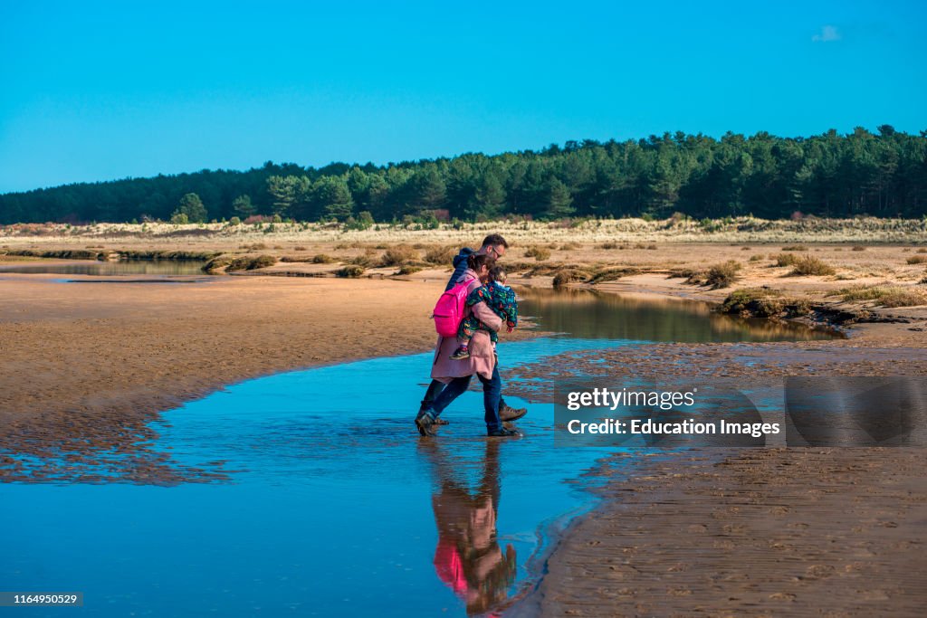 A family cross water pools left by the tide on sandy beach at Holkham bay, North Norfolk coast, East Anglia, England, UK