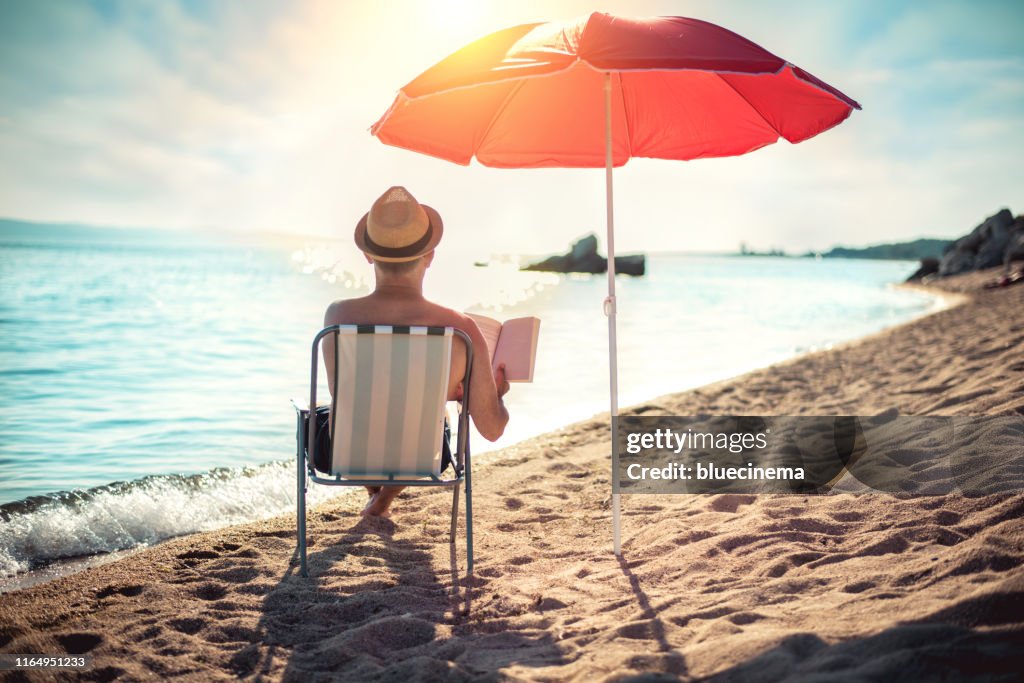 Uomo che legge il libro sulla sedia a sdraio sotto l'ombrellone vicino al mare