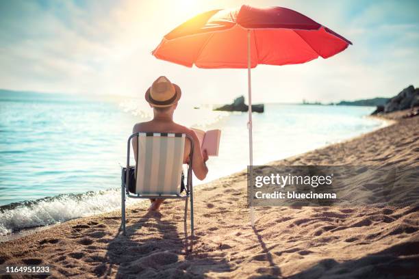 man läser bok i solstol under solparasoll nära havet - book blue bildbanksfoton och bilder