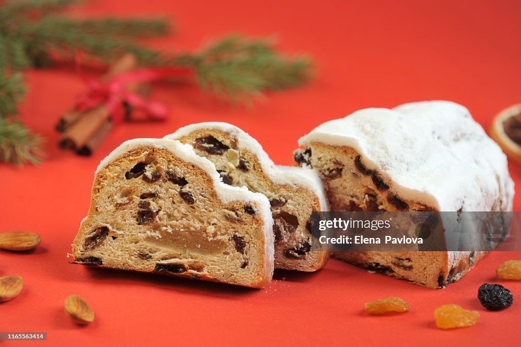 Christmas stollen on a red background. The frame space is occupied by slices of cake, cinnamon sticks, raisins and almond nuts. Close-up.