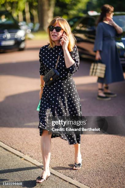 Guest wears sunglasses, a black bag, a black dress with white polka dots, black sandals with bow, outside Miu Miu Club 2020, on June 29, 2019 in...