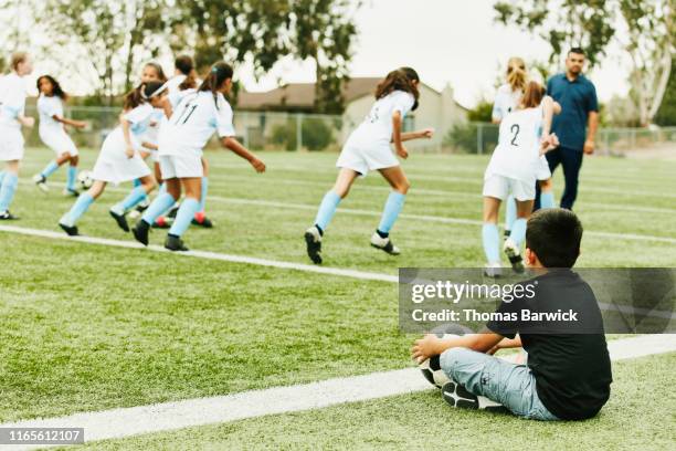 young boy sitting on sidelines watching sisters soccer team warm up for game - african american girl wearing a white shirt stock pictures, royalty-free photos & images