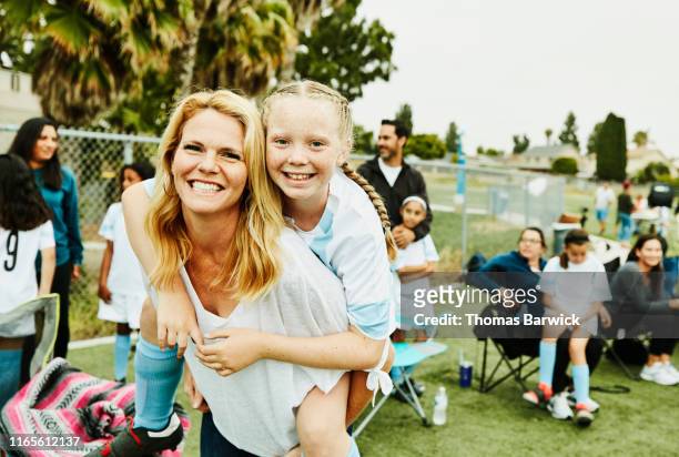 smiling mother giving daughter piggy back ride on sidelines after soccer game - familie sport stock pictures, royalty-free photos & images