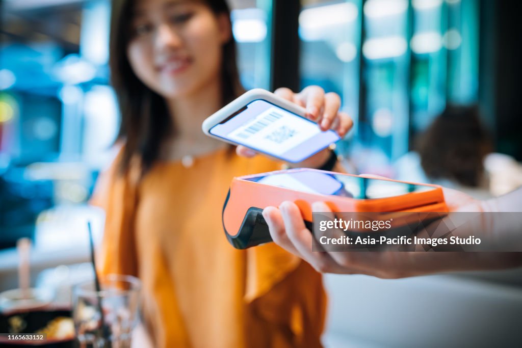 Mujer joven asiática pagando con teléfono inteligente en un café.