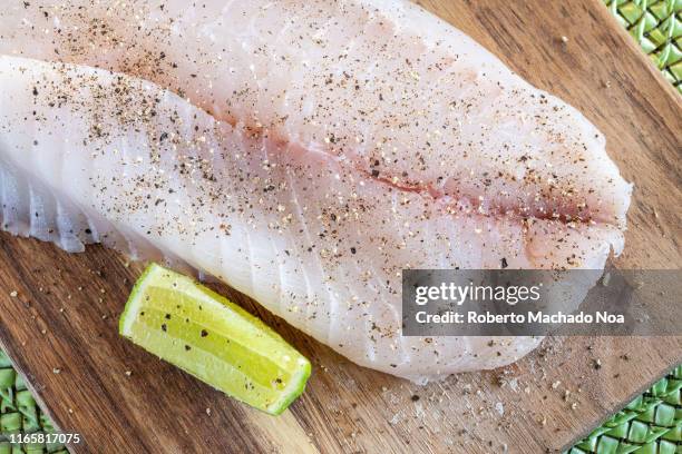 Raw tilapia fillet over a wooden cutting board in the kitchen. The fish is marinated with salt and pepper. There is a wedge of lime by the side.