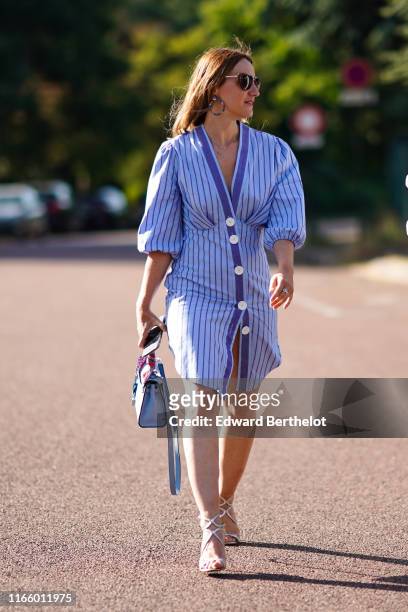 Guest wears sunglasses, a blue striped dress with buttons, a blue bag, earrings, shoes, outside Miu Miu Club 2020, on June 29, 2019 in Paris, France.