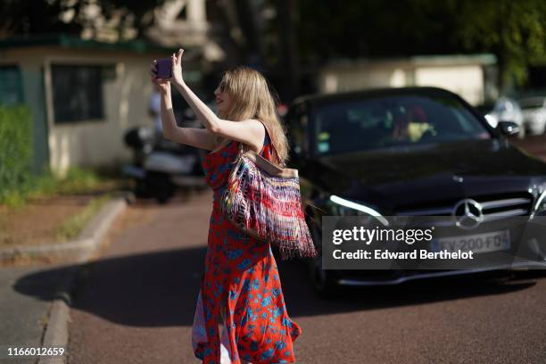 Guest wears a multicolor fringed bag, a red and blue floral print dress, takes a picture with a smartphone, outside Miu Miu Club 2020, on June 29,...