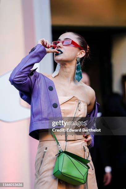 Guest wears rhinestone embellished red sunglasses, mirror earrings, a mauve leather jacket, a peach-color leather off-the-shoulder dress, a green bag...