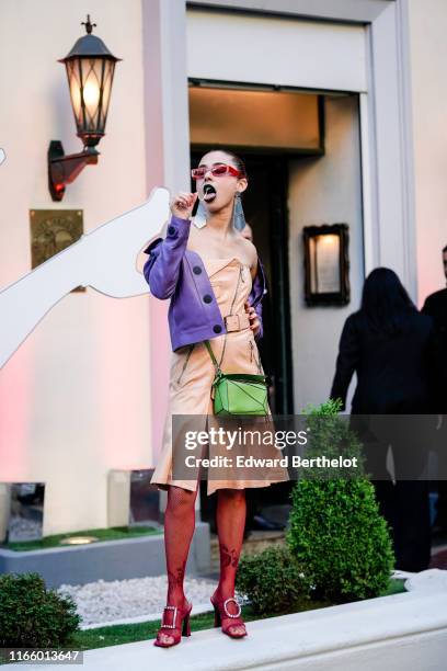 Guest wears rhinestone embellished red sunglasses, mirror earrings, a mauve leather jacket, a peach-color leather off-the-shoulder dress, a green...