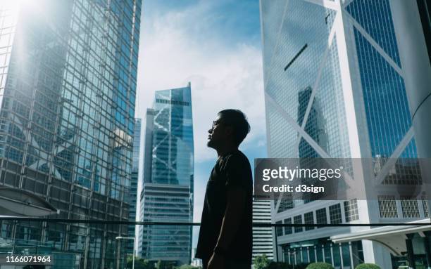 silhouette of professional young businessman standing against contemporary financial skyscrapers and looking up into sky with positive emotion - ergens overheen kijken stockfoto's en -beelden