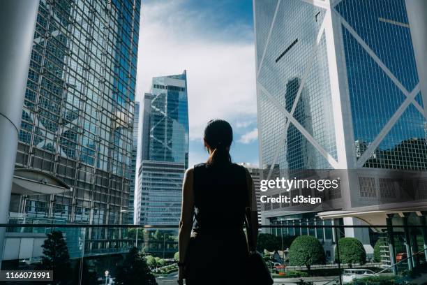 rear view of professional young businesswoman standing against contemporary financial skyscrapers in downtown financial district and looking up into sky with positive emotion - business professional ストックフォトと画像