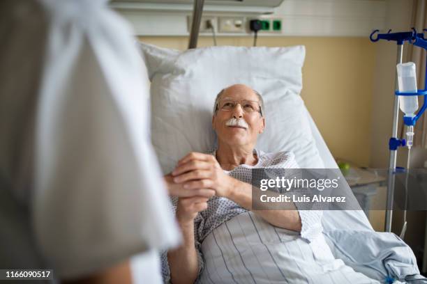 senior man holding hand of female nurse in hospital room - health care professional with patient stock-fotos und bilder