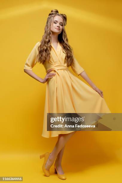 a cute young girl in a yellow dress, in shoes with heels on a yellow background with a haircut and curly long hair. yellow fashionable, stylish, youth clothing. salon hairstyles and makeup. - robe jaune photos et images de collection