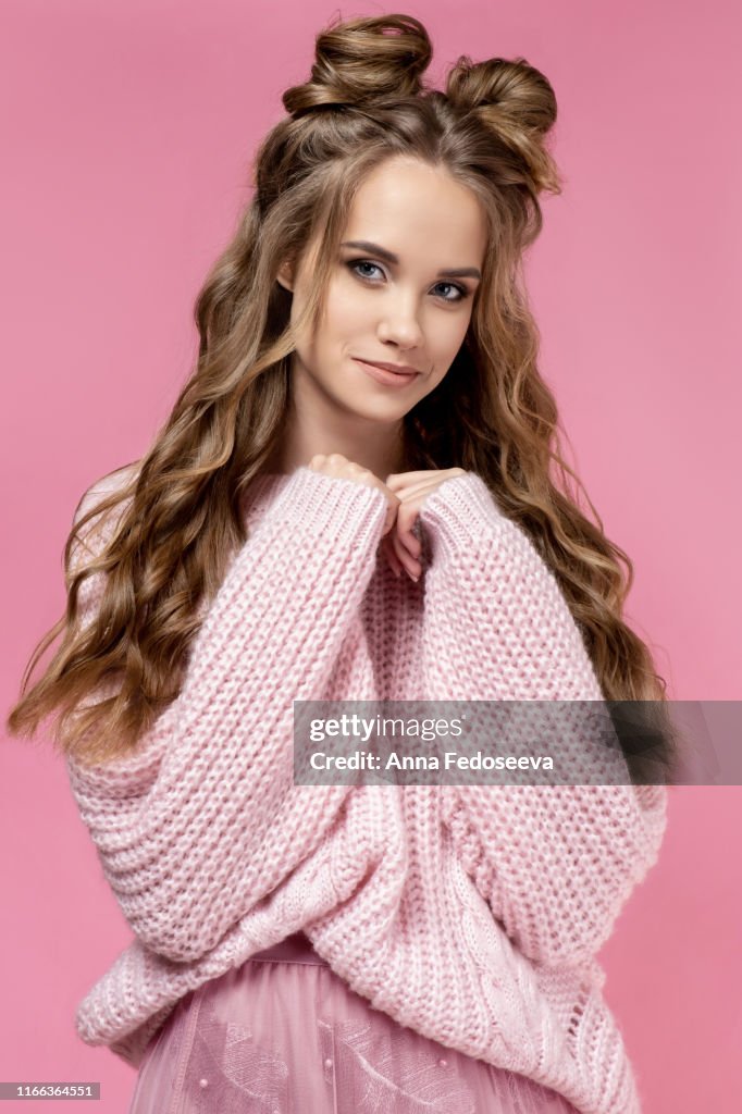 Pretty young girl in a pink sweater on a pink background with a haircut and curly long hair. Pink total look. Fashionable, stylish, youth clothing. Salon hairstyles, women barbershop and makeup.