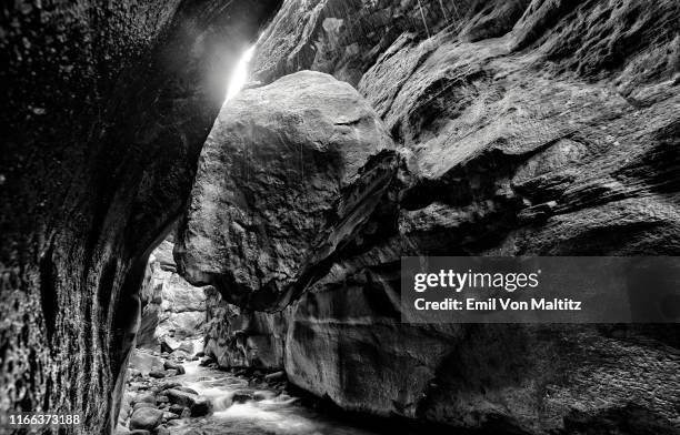 the massive boulder that is wedged between the two rock faces of the rainbow gorge corridor. a stream is flowing below the suspended boulder while water rains down from above. cathedral peak, drakensberg, kwazulu natal, south africa. black and white image - cathedral imagens e fotografias de stock