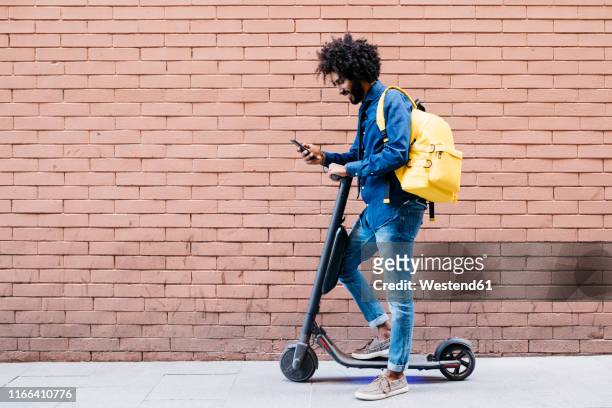 young man with backpack and e-scooter standing in front of brick wall looking at cell phone - on the move stock pictures, royalty-free photos & images