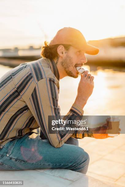 tattooed man with coffee to go eating  doughnut at sunset - donut man stock pictures, royalty-free photos & images