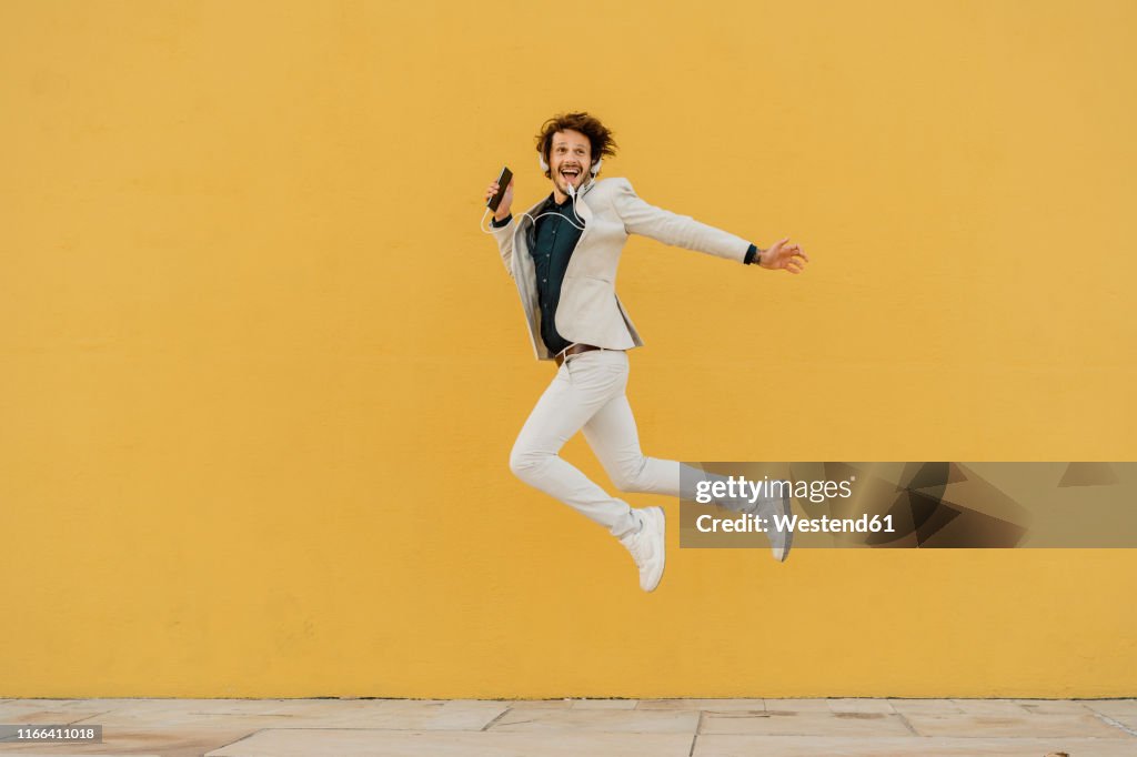 Happy businessman jumping in the air in front of yellow wall listening music with headphones