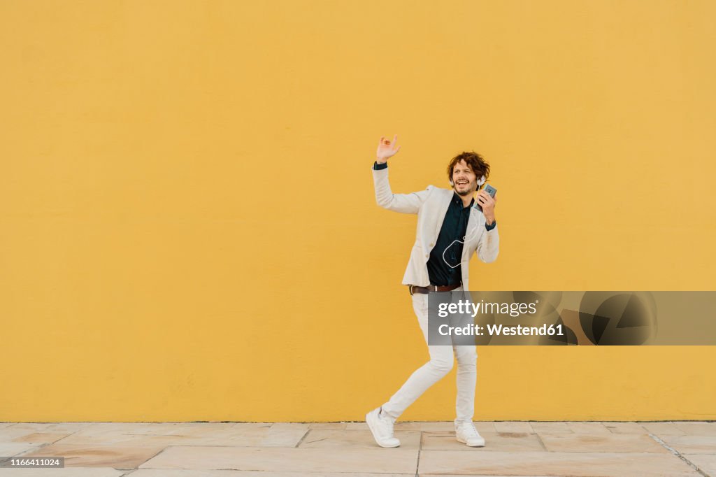 Businessman singing and dancing in front of yellow wall listening music with headphones and smartphone