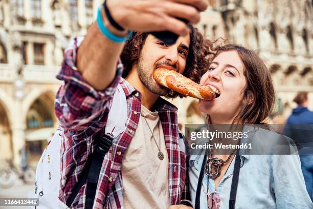 young couple taking a selfie with brezel in the mouth, munich, germany - funny selfie stockfoto's en -beelden