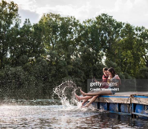 young couple having a drink and splashing with water on jetty at a remote lake - couple jetty stock pictures, royalty-free photos & images