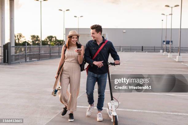 happy young couple with cell phone, longboard and electric scooter on parking deck - roller skates stock-fotos und bilder