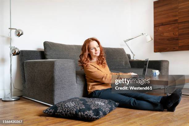 portrait of smiling redheaded woman sitting on floor in the living room using laptop - design laptop woman stock-fotos und bilder