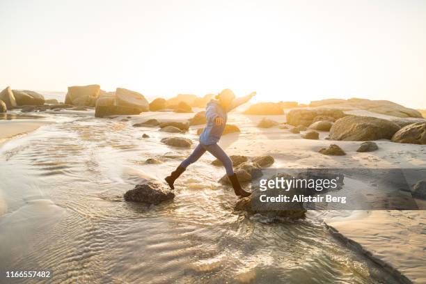 senior woman stepping across stream on the beach at sunset - überqueren stock-fotos und bilder