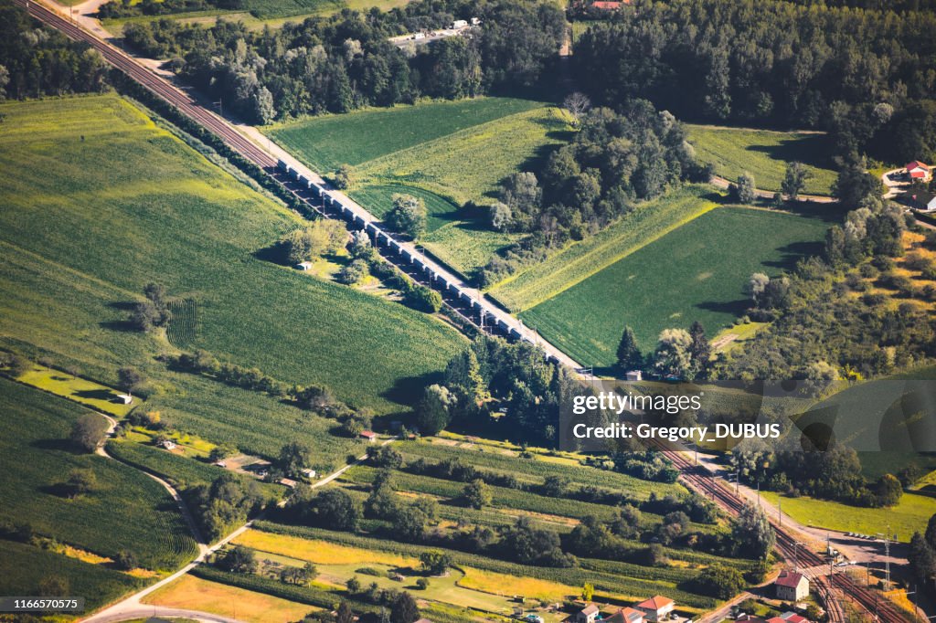 Aerial view on freight train railroad track crossing in diagonal the French countryside in summer, in middle of vibrant green fields from Ain department, Auvergne-Rhone-Alpes region