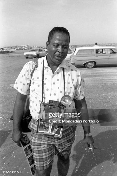 Portrait of a man with a Polaroid Land camera 104 around his neck holding some instant photos. United States, 1969